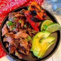 Carne Asada on a black skillet with sliced avocado, roasted peppers, and lime wedges