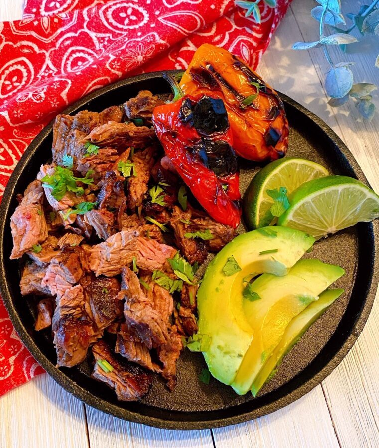 Carne Asada on a black skillet with sliced avocado, roasted peppers, and lime wedges