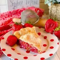 Big slice of Strawberry Rhubarb Pie on a fun polka dot plate with fresh strawberries surrounding it and a pretty background.