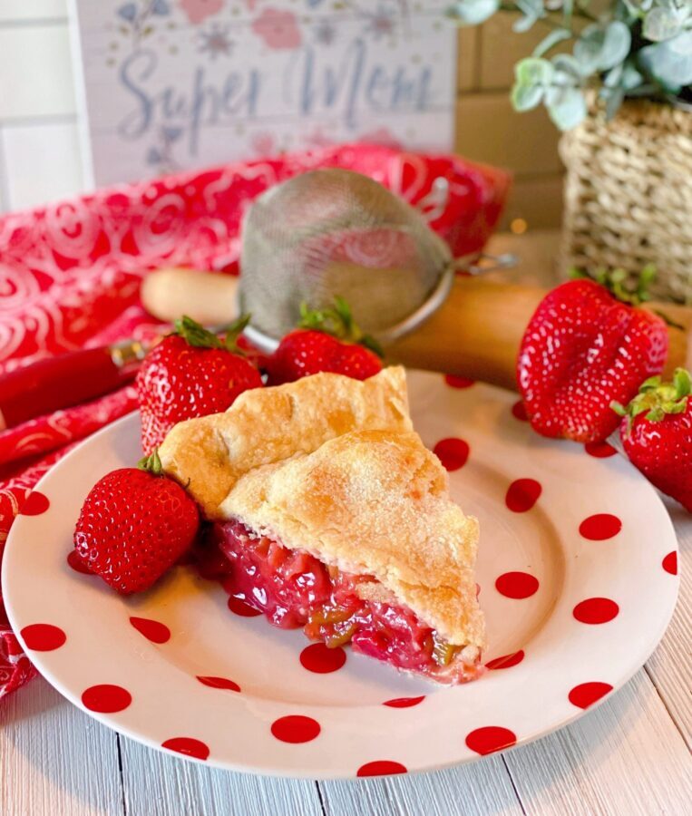 Big slice of Strawberry Rhubarb Pie on a fun polka dot plate with fresh strawberries surrounding it and a pretty background.