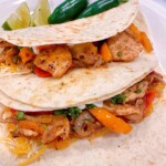 A couple of chicken fajitas made up in flour tortillas on a white plate with peppers and lime.