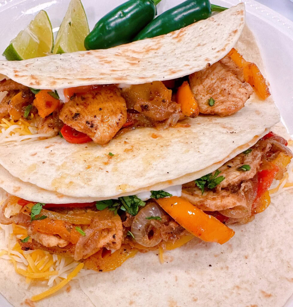 A couple of chicken fajitas made up in flour tortillas on a white plate with peppers and lime.