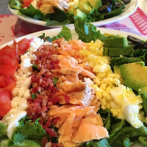 Cobb Salmon Salad with rows of grilled salmon, bacon bits, diced hard boiled eggs, sliced avocado, and diced tomatoes on a bed of leafy greens.