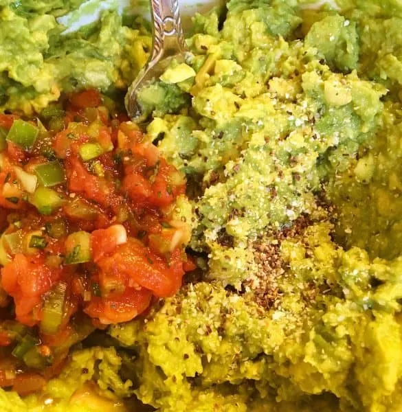 Mashed Haas Avocados with pepper, garlic salt, and a scoop of fresh salsa being mixed together. These ingredients create the best homemade guacamole.