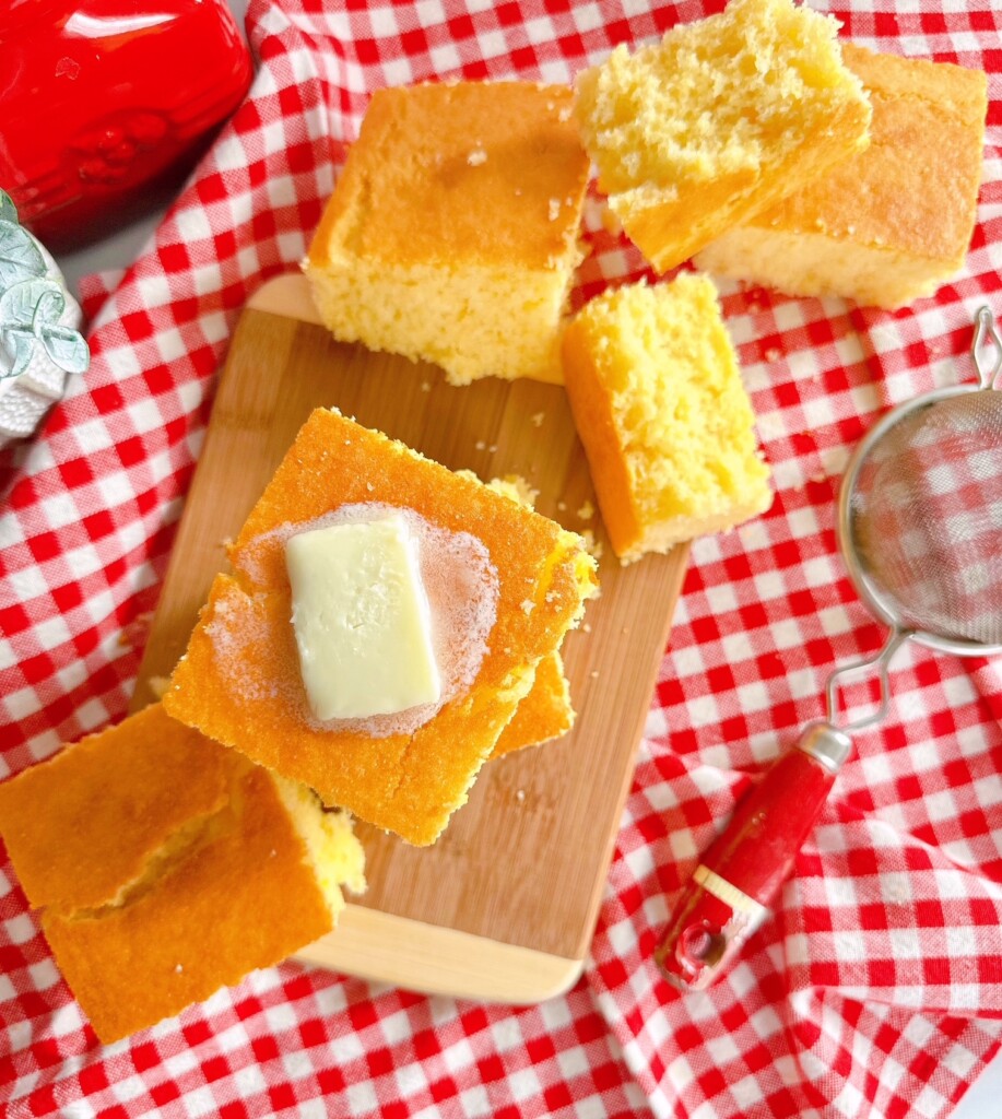 Buttery Sweet Cornbread cut and on a wooden board with a checked tablecloth underneath it.