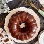 Overhead photo looking down on Chocolate Cake with Satin drizzle, surrounded by broken chocolate pieces and a wire whisk.