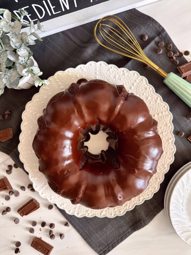 Overhead photo looking down on Chocolate Cake with Satin drizzle, surrounded by broken chocolate pieces and a wire whisk.