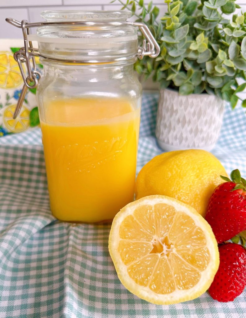 Homemade microwave lemon curd in a mason jar surrounded by lemons and fresh strawberries.