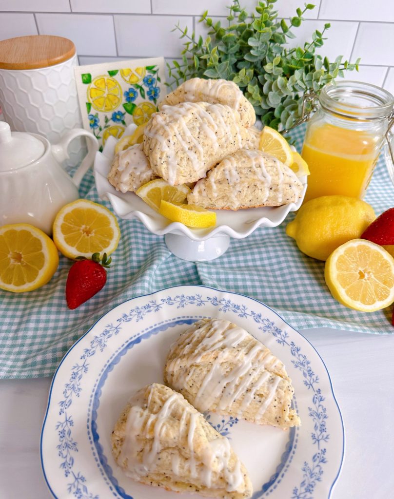 Lemon Poppy Seed Scones on a cake plate and extra's on a serving plate with lemons, strawberries, and fresh lemon curd.