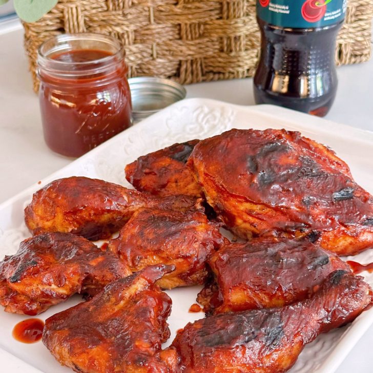 Perfectly grilled Cherry Dr Pepper Chicken on a white platter with more BBQ sauce in the background.