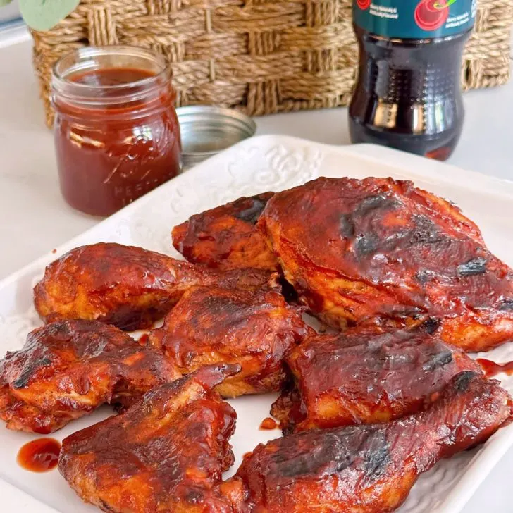 Perfectly grilled Cherry Dr Pepper Chicken on a white platter with more BBQ sauce in the background.
