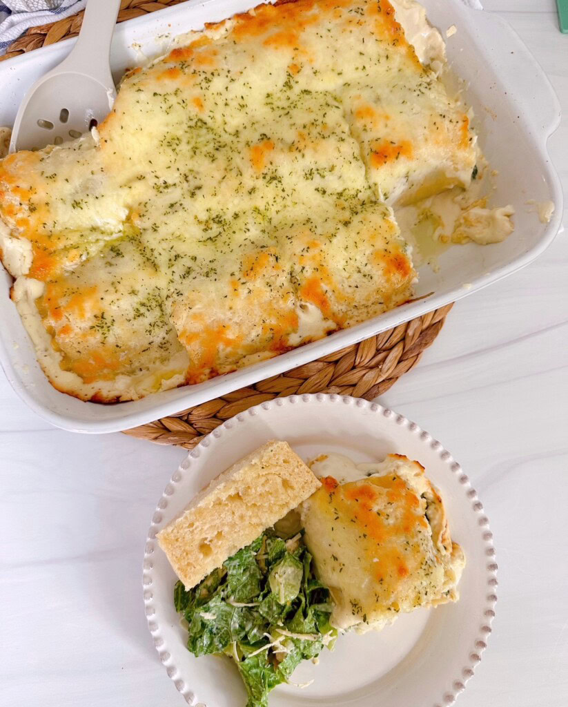 Overhead shot of the casserole dish filled with Spinach Alfredo Chicken Lasagna Roll-ups and a plate filled with a serving of same, salad, and garlic bread.