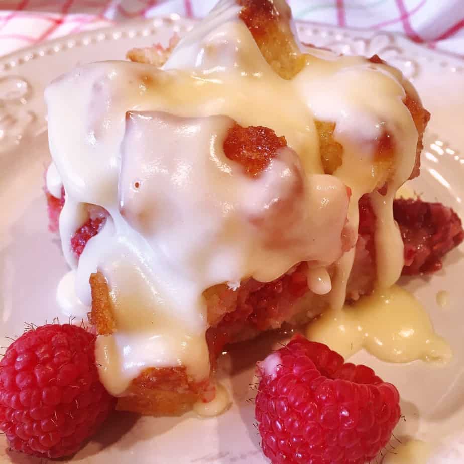 Square Piece of Raspberry Bread Pudding with Creamy Vanilla Cream Sauce poured over the top and fresh raspberry garnish on a white plate.