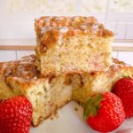 Rhubarb Sour Cream Coffee Cake cut into squares and stacked on a cake plate with fresh strawberries around it.