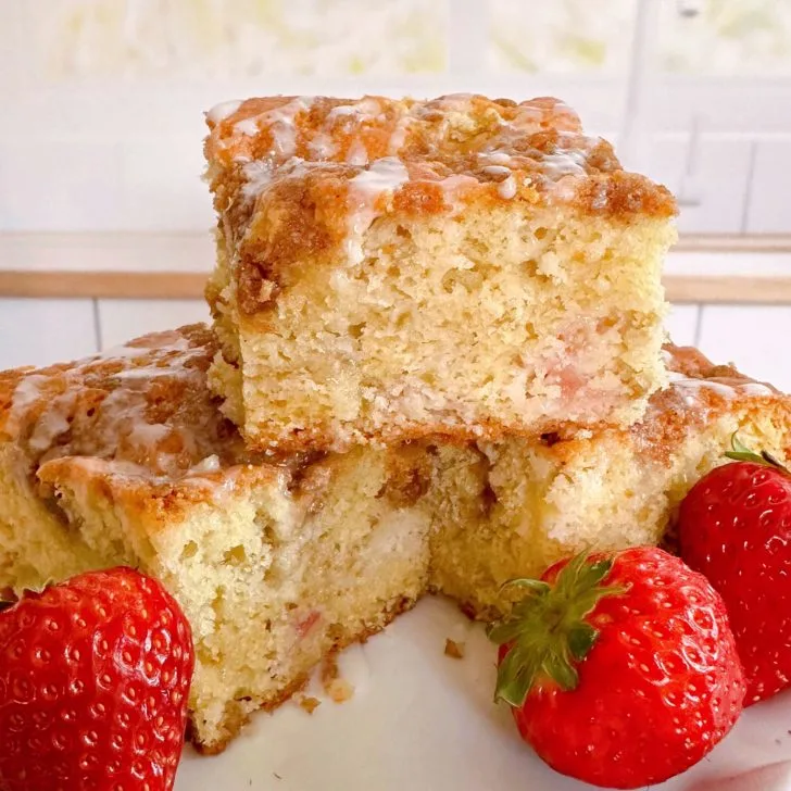 Rhubarb Sour Cream Coffee Cake cut into squares and stacked on a cake plate with fresh strawberries around it.