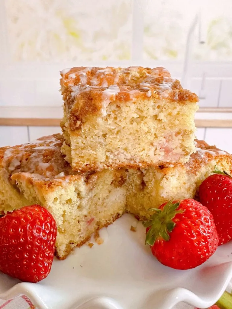 Rhubarb Sour Cream Coffee Cake cut into squares and stacked on a cake plate with fresh strawberries around it.