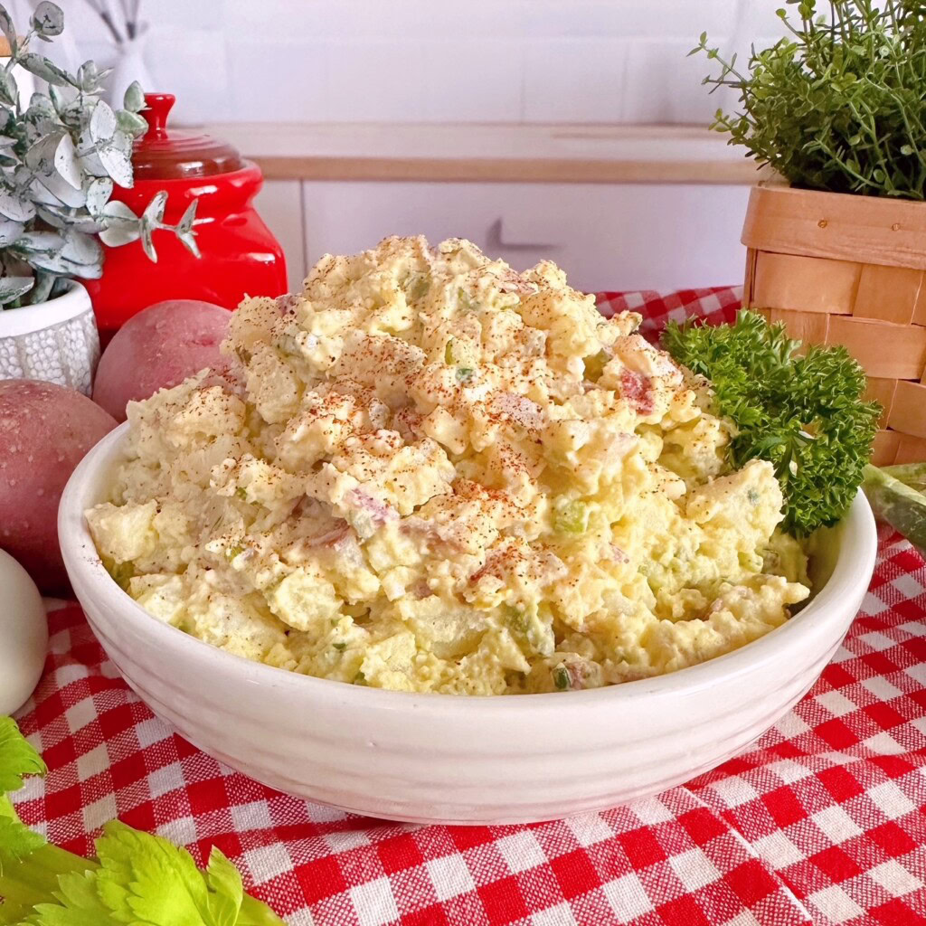 White serving bowl full of classic red potato salad with ingredients to make it surrounding it.