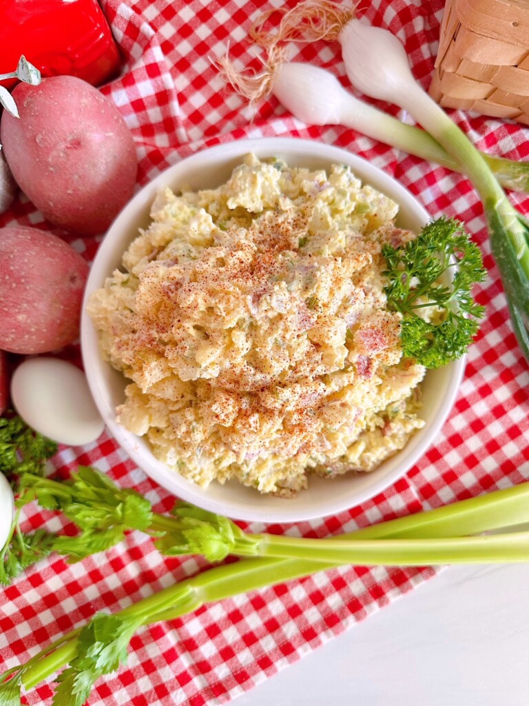 Classic Red Potato Salad with all the ingredients surrounding it in a white serving bowl on a red and white checkered cloth.