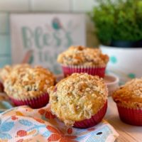 Pile of Mixed Berry Muffins on the table with plants and a bless this nest sign in the background