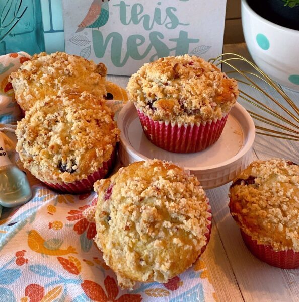 Pile of Streusel Pecan topped berry muffins on a table with a wire whisk and polka dot vase with a plant in the background