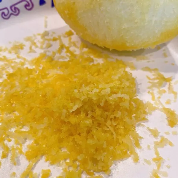 Zest of lemon on a paper plate with the zested lemon in the background.