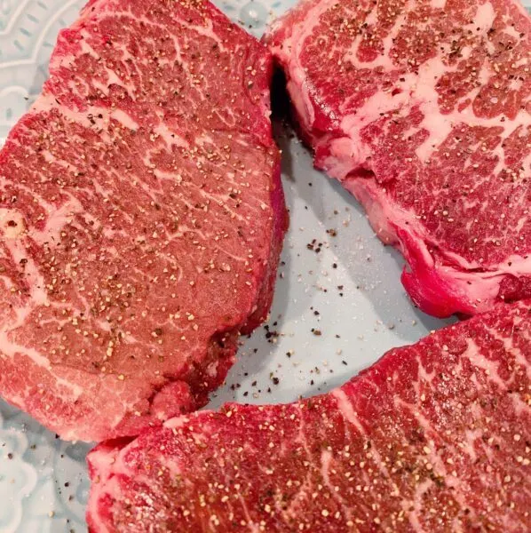 Raw Denver Steaks, three of them, on a plate seasoned with Salt and Pepper and resting to warm up before cooking.