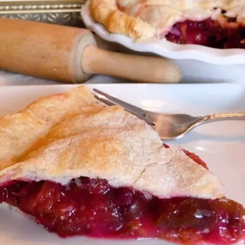 Big slice of Plum Pie with a fork ready to eat. Rolling Pin and Whole Plum Pie in the background