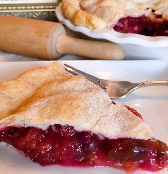 Big slice of Plum Pie with a fork ready to eat. Rolling Pin and Whole Plum Pie in the background