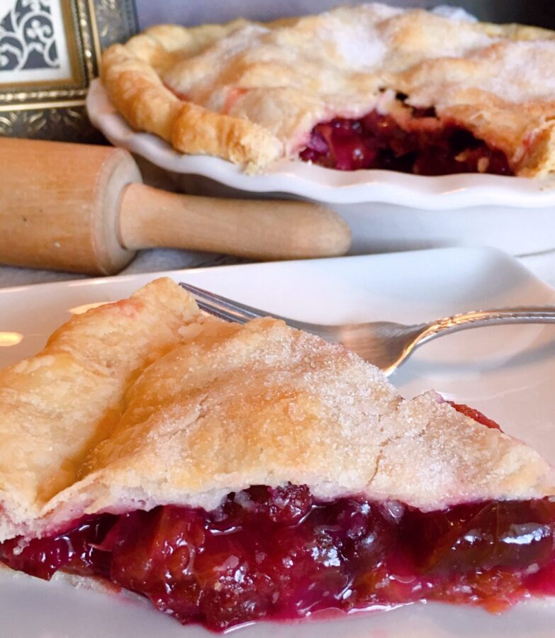 Slice of Plum Pie with whole pie in the background and a rolling pin on the kitchen counter