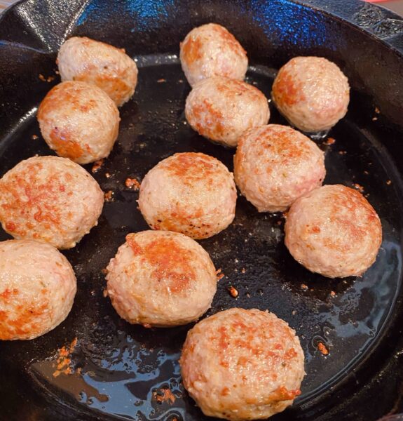 Large cast iron skillet on a stove top with browning meatballs.