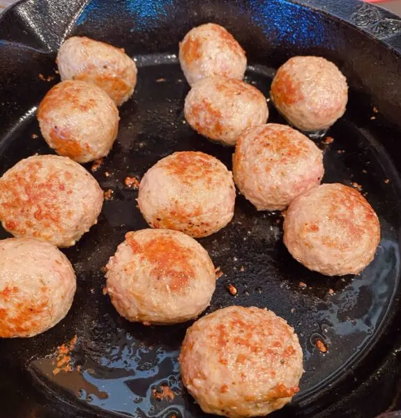 Large cast iron skillet on a stove top with browning meatballs.