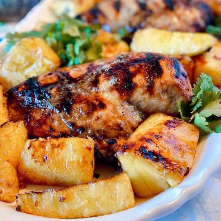 Close-up photo of a grilled jerk chicken breast on a white platter with grilled pineapple spears.