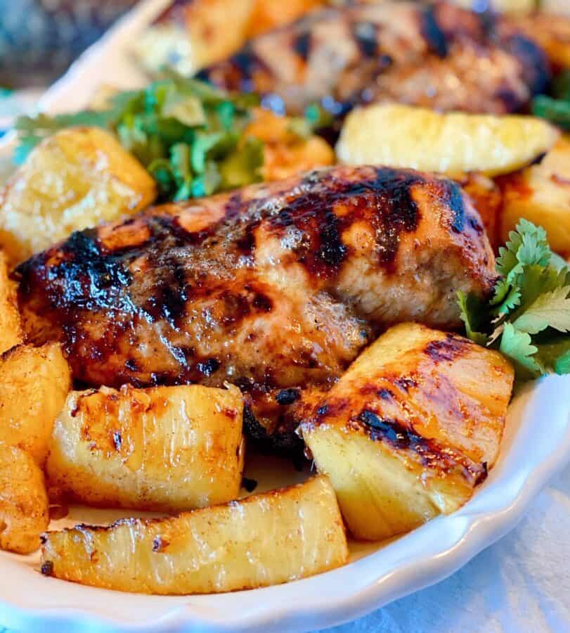 Close-up photo of a grilled jerk chicken breast on a white platter with grilled pineapple spears.