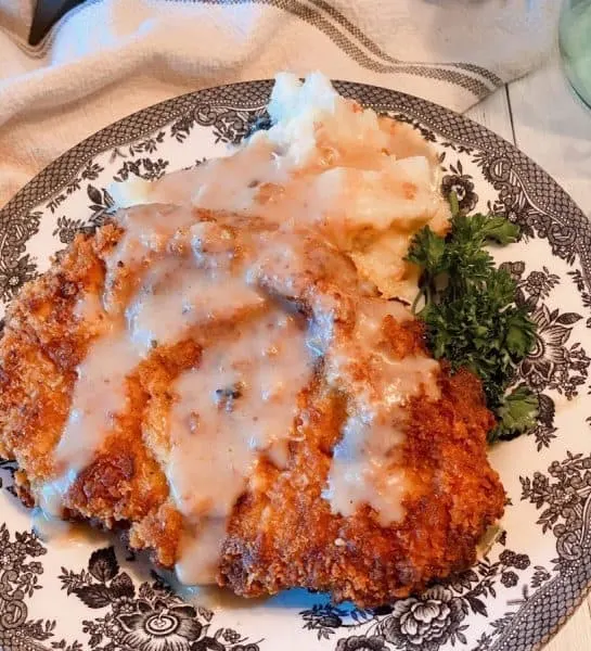 Chicken Fried Pork Steak on a bed of mashed potatoes with creamy country gravy on a plate with a garnish of parsley.