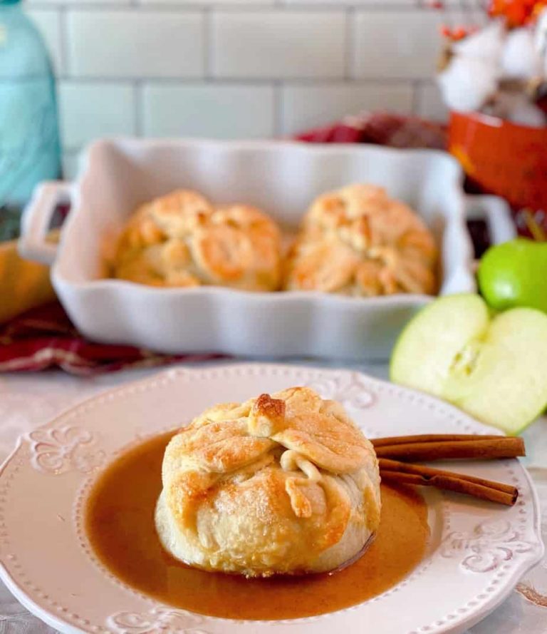Country Apple Dumpling on a plate with cinnamon sauce and more apple dumplings in a baking dish in the background
