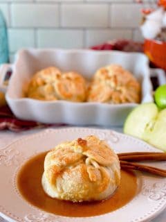 Country Apple Dumpling on a plate with cinnamon sauce and more apple dumplings in a baking dish in the background