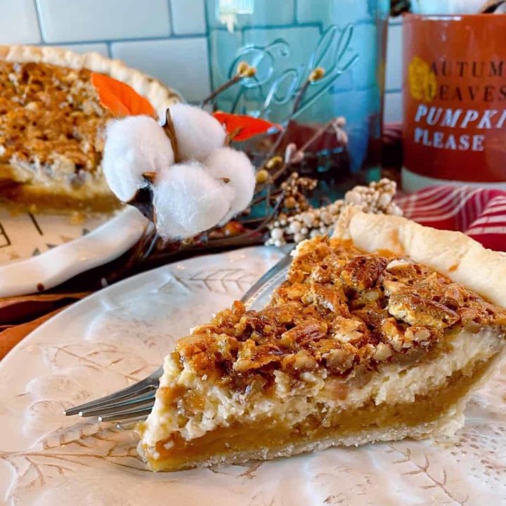 Slice of Caramel Pecan Cheesecake Pie on a tan plate with a fork and whole pie in the background with fall decor.