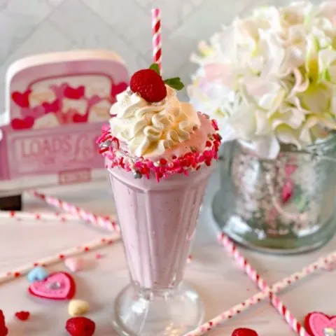 White Chocolate Raspberry Milk Shake in an old fashioned glass rimmed with valentine sprinkles, a festive striped straw, and valentines candies.