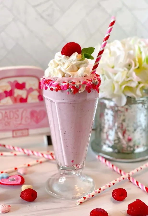 White Chocolate Raspberry Milk Shake in an old fashioned milk shake glass rimmed with valentine sprinkles, topped off with a cloud of whipped cream and a fresh raspberry and fun striped straw.
