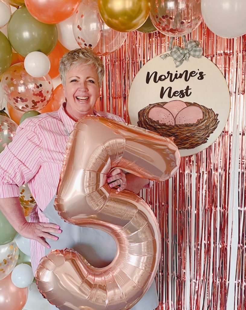 Norine holding a large number 5 balloon with a big balloon arch behind her and her logo.