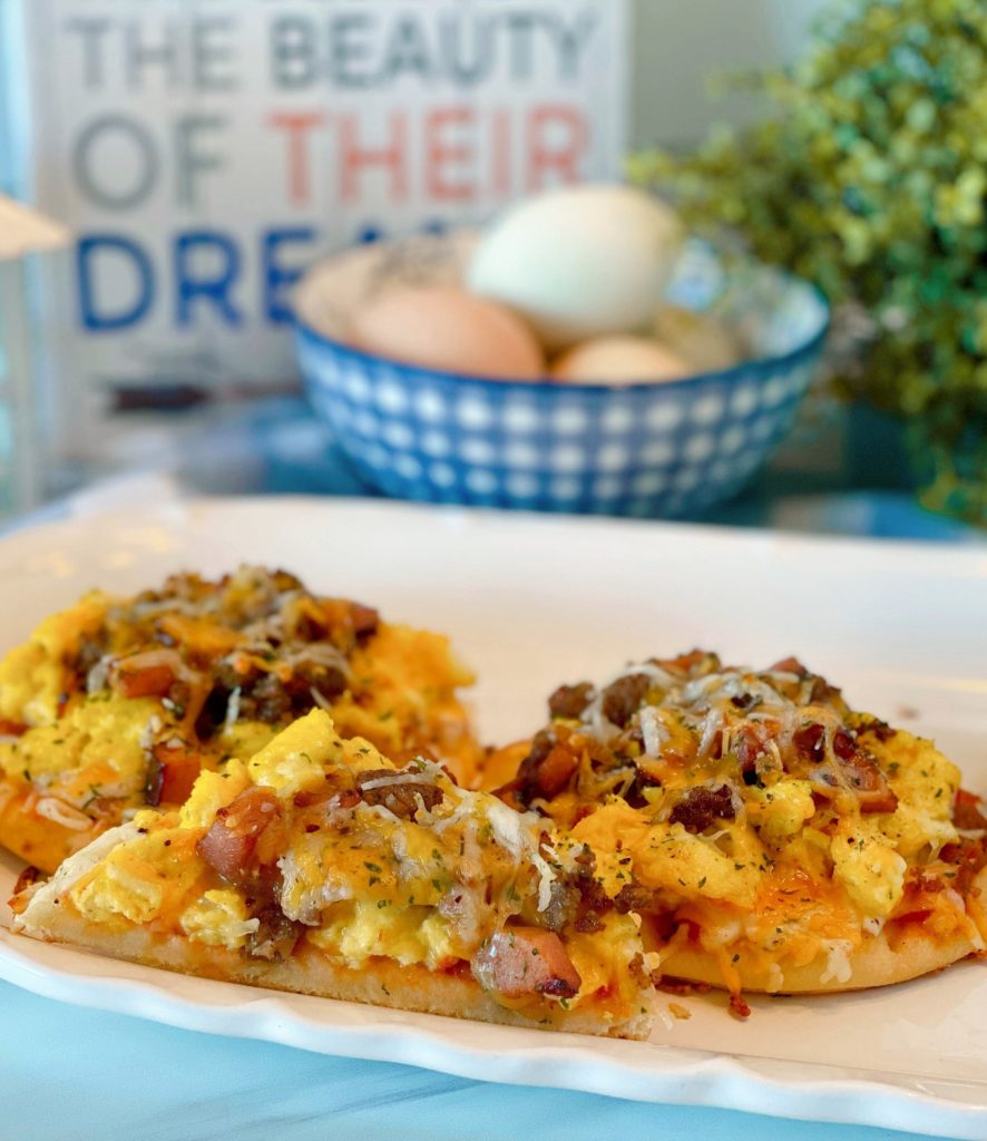 Individual Naan Breakfast Pizzas on a white plate with eggs in a bowl in the background. One pizza is cut in half revealing the toppings inside.