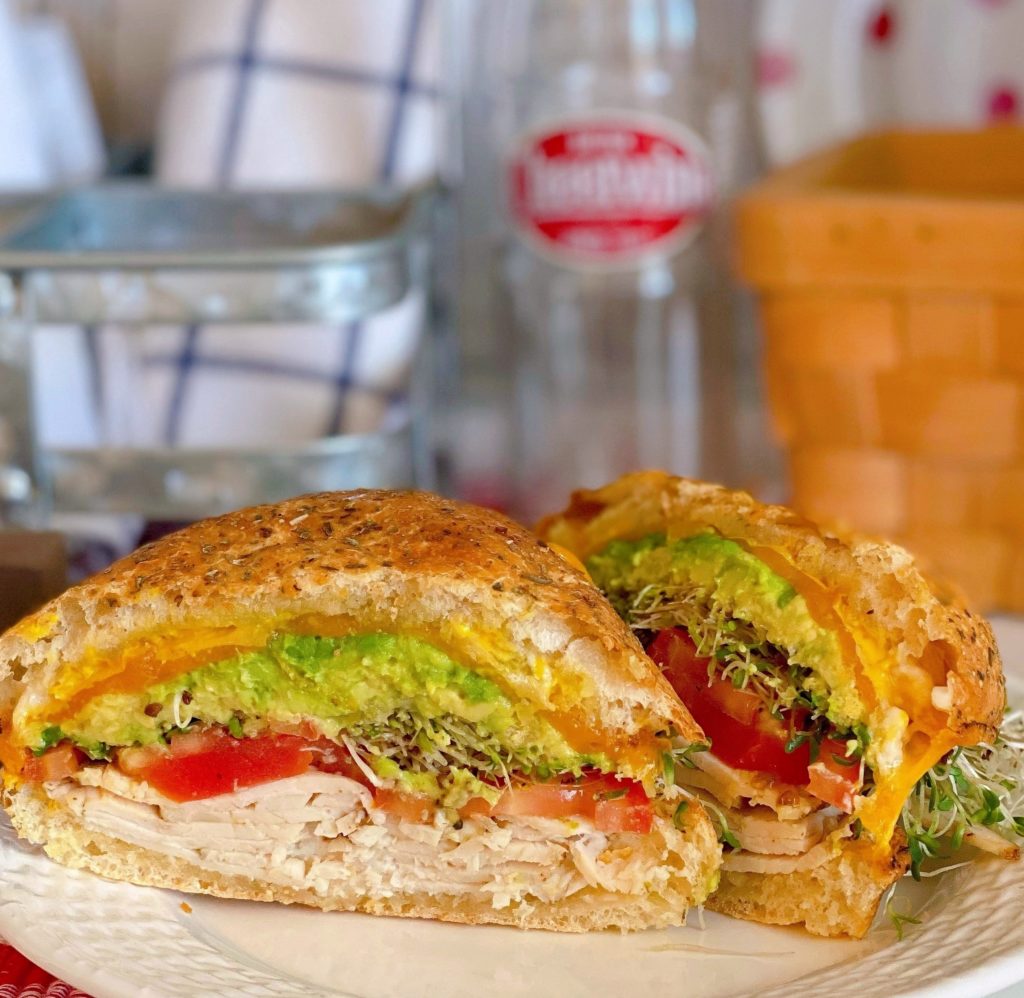 Sliced Air Fryer Toasted Turkey Sandwich with lettuce, tomatoes, avocado, sprouts, melted cheese on a plate with a soda bottle in the back ground.