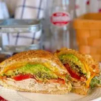 Sliced Air Fryer Toasted Turkey Sandwich with lettuce, tomatoes, avocado, sprouts, melted cheese on a plate with a soda bottle in the back ground.