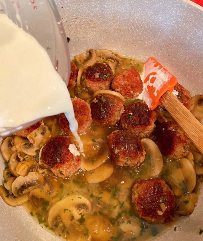 Adding cream mixture to meatball mixture in pan on stove.