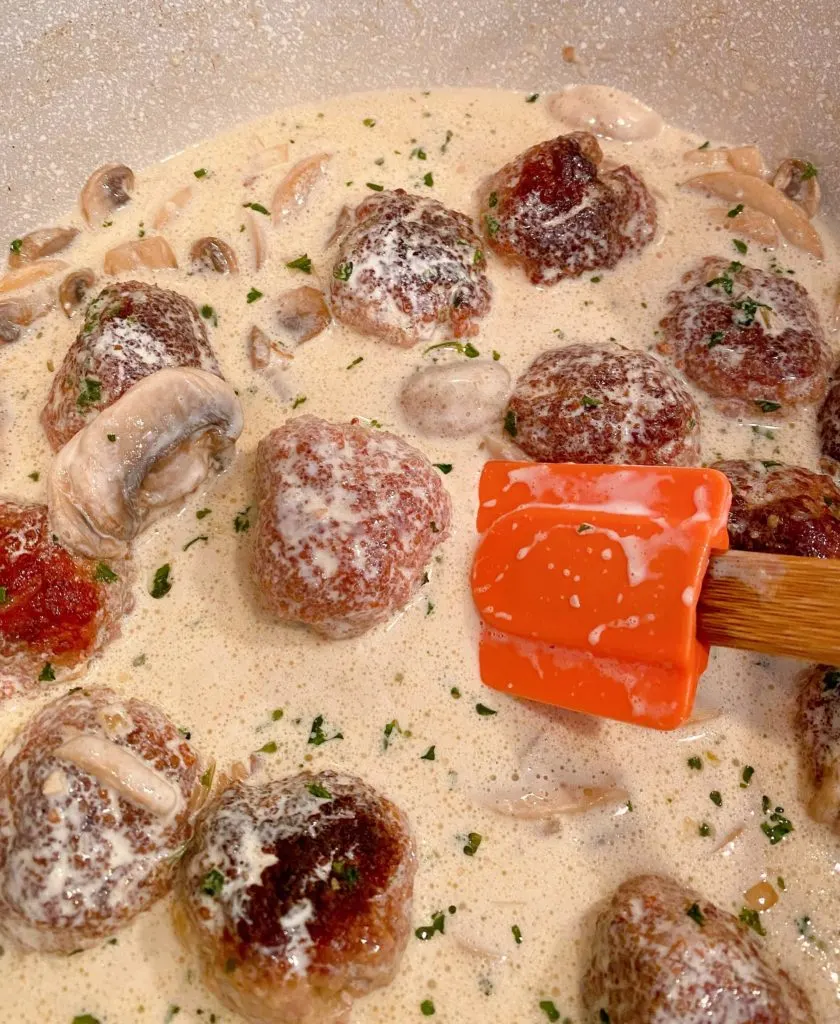 Stirring cream into meatballs with orange spatula.