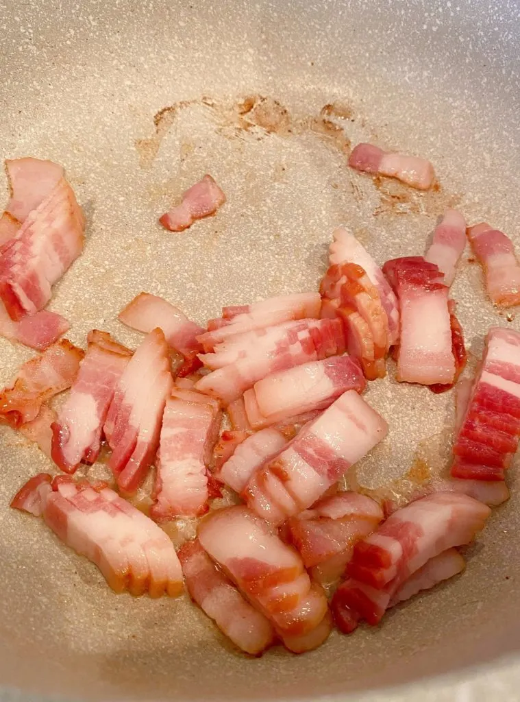 Bite size piece of bacon browning in a skillet.
