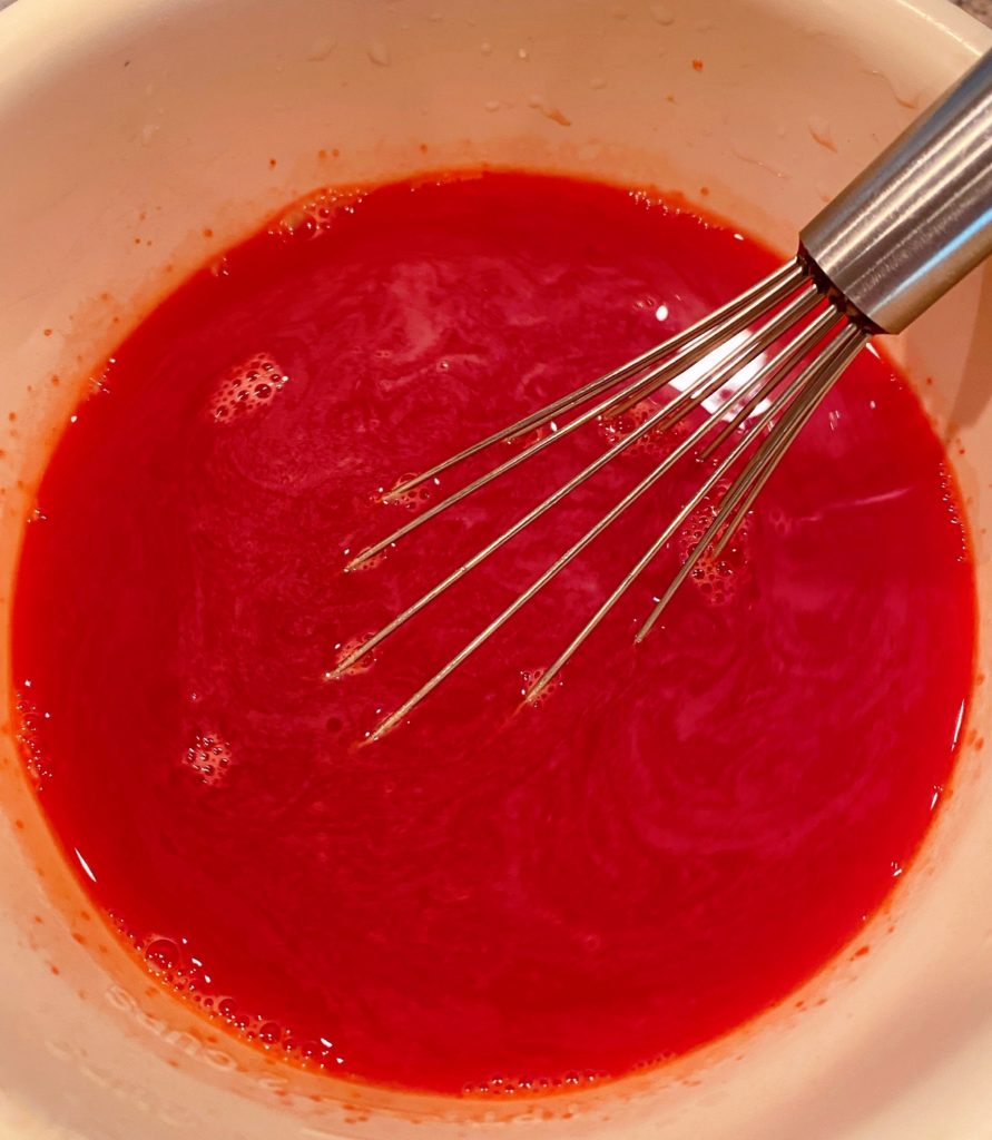 Small plastic bowl with raspberry jello and water and a wire whisk mixing until combined.