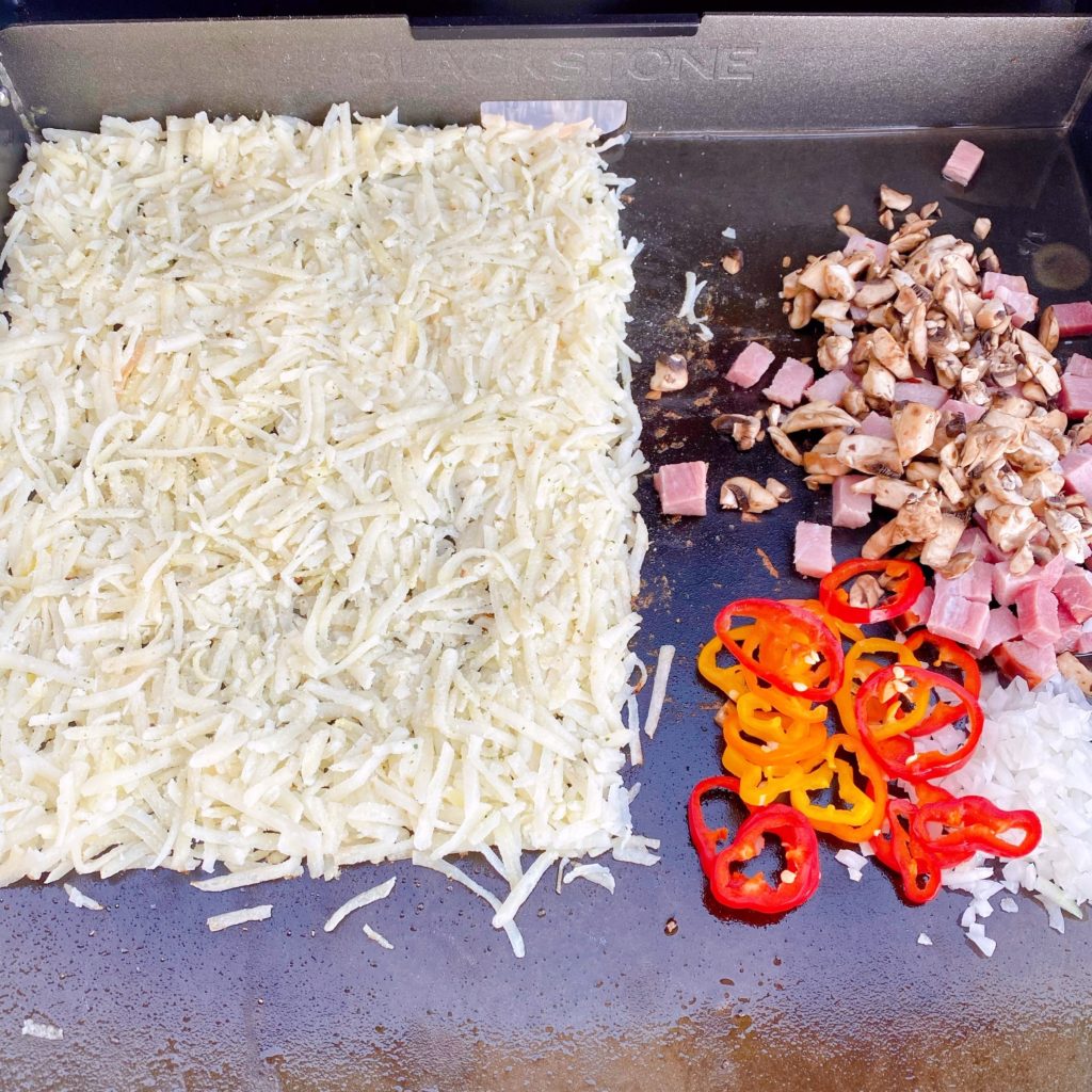 Hash Browns spread out on the griddle. Vegetables on the other side cooking.