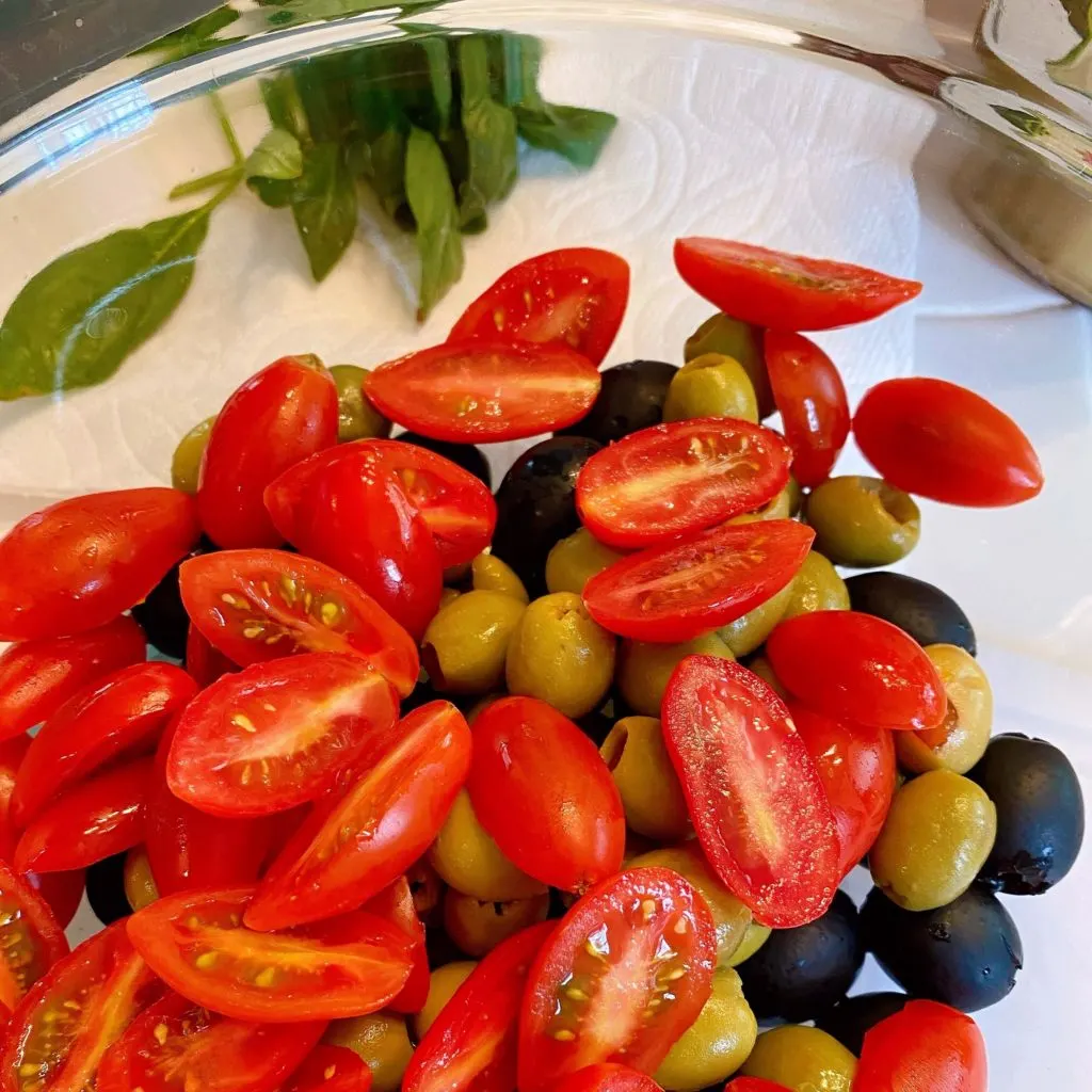Adding sliced grape tomatoes to olives in large bowl.