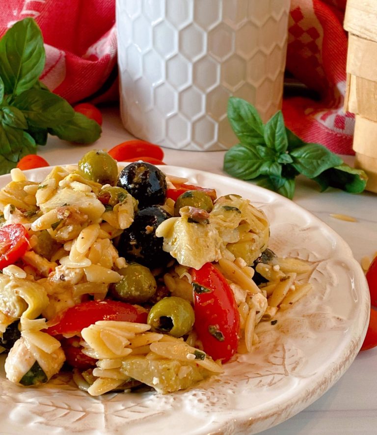 Muffuletta Olive Pasta Salad close up photo on a white plate with fresh basil surrounding it.